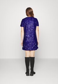 Kortärmad klänning ovanför knäet täckt av lila paljetter; kombinerad med knähöga svarta strumpor och platåskor.