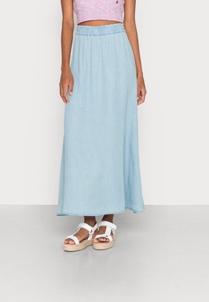 Femme portant une jupe longue bleu clair avec une taille élastique, associée à des sandales à plateforme blanches et un top court rose.