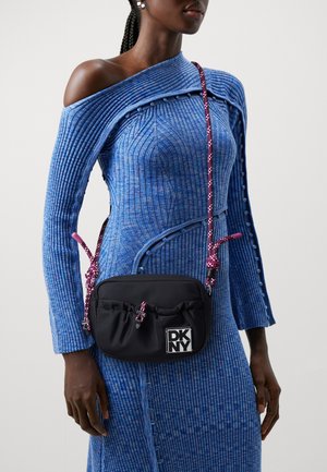 Sac à bandoulière noir DKNY avec une sangle en corde rose porté sur une robe bleue à côtes, à une épaule avec des détails de boutons, montré sur une femme de la moitié du torse vers le haut.