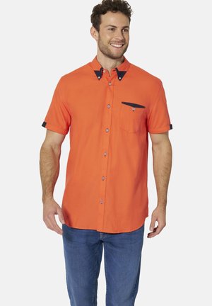 Mann lächelnd, trägt ein leuchtend orangefarbenes kurzärmeliges Hemd mit dunklem Kragen und Taschen-Details, kombiniert mit blauen Jeans.