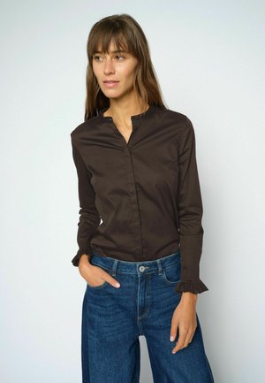 Frau mit glatten braunen Haaren, die eine dunkelbraune Langarmbluse und blaue Jeans trägt, steht mit einer Hand in der Tasche.