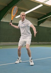 Hvit tennis skjorte med blå detaljer, hvite shorts og svarte og hvite tennissko. Spilleren holder en oransje tennisracket på en blå bane.