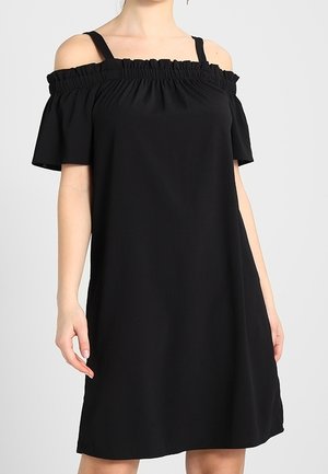 Kvinde iført en sort off-shoulder kjole med flæsekant og korte ærmer mod en ensfarvet lys baggrund.