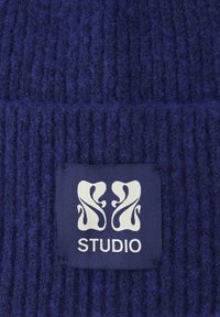 Námořnicky modrá žebrovaná pletená látka s texturovaným vzhledem. Má bílou etiketu s abstraktním designem a nápisem "STUDIO".