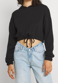 Hoodie cropped nero con vita regolabile con stringa, maniche lunghe e polsini elasticizzati, abbinato a jeans a vita alta blu chiaro. Tessuto dalla trama liscia.