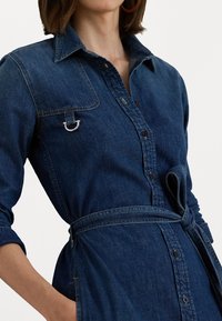 Abito camicia in denim blu scuro con bottoni frontali, colletto e cintura legata in vita. Presenta un dettaglio con anello in metallo argentato sulla tasca del petto.