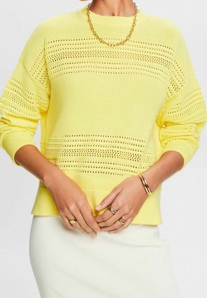 Personne portant un pull en maille jaune avec des rayures ajourées horizontales, un collier chaîne en or, des bagues, des bracelets et une jupe blanche.