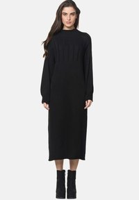 Czarna dzianinowa sukienka z prążkowanym wzorem, długimi rękawami, wysokim kołnierzem i prostą sylwetką; w zestawieniu z czarnymi botkami za kostkę.