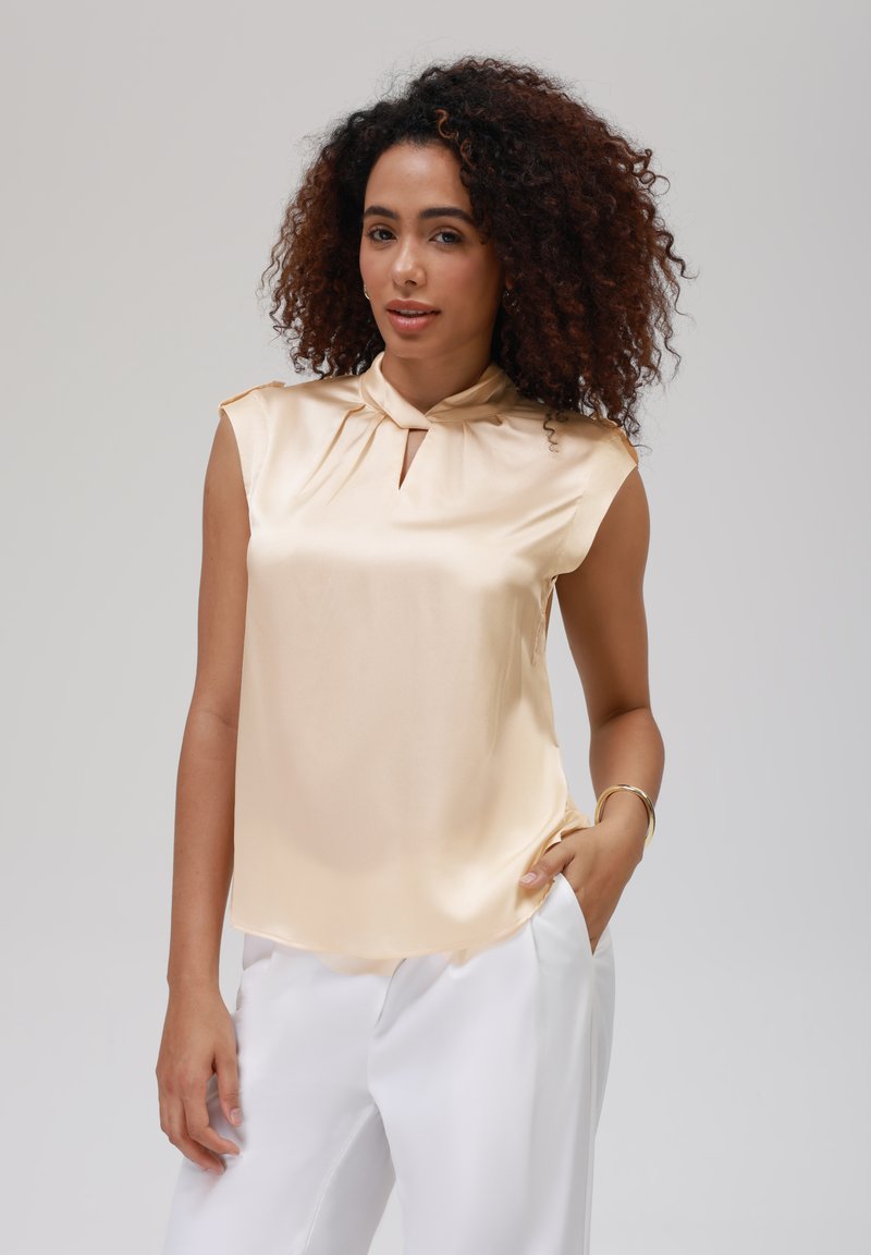 Blouse sans manches en satin de couleur or clair avec un pli au niveau du cou et un détail en trou de serrure, texture lisse et coupe décontractée, associée à un pantalon blanc.
