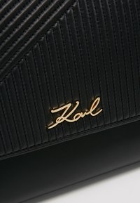 Accessoire en cuir noir avec des coutures verticales et un détail en métal doré représentant la signature "Karl" sur la partie inférieure lisse.