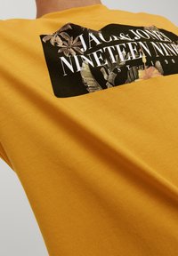 Uomo che indossa una maglietta gialla senape con un grafico rettangolare nero che presenta un testo bianco e un design di foglie tropicali sul retro.