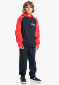 Hoodie en polaire bleu marine et rouge avec poche frontale, arborant un logo "Quiksilver" en relief. Associé à un pantalon de jogging bleu marine assorti et des chaussures noires à enfiler.