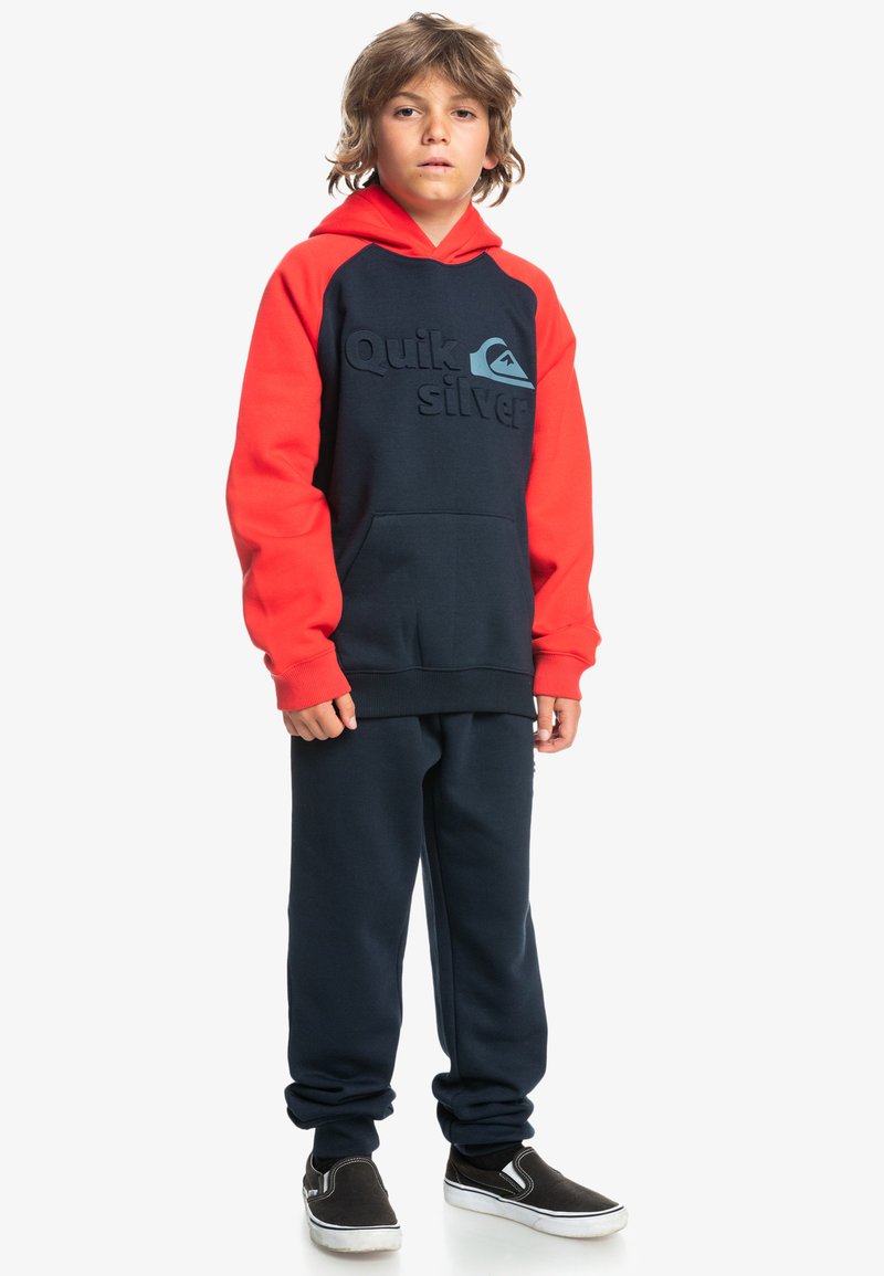 Hoodie en polaire bleu marine et rouge avec poche frontale, arborant un logo "Quiksilver" en relief. Associé à un pantalon de jogging bleu marine assorti et des chaussures noires à enfiler.