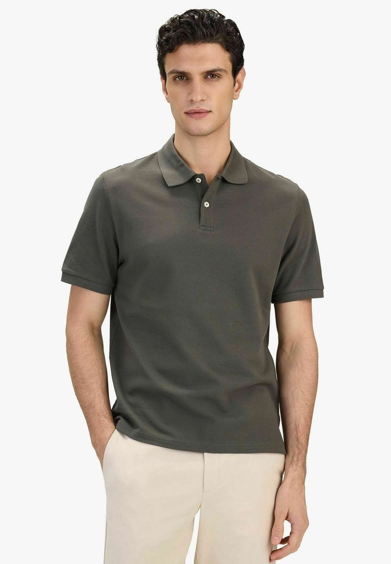 Homme aux cheveux bouclés foncés portant un polo à manches courtes vert foncé et un pantalon beige, debout avec une main dans la poche.