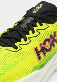 Baskets de course jaunes vives avec lacets et accents noirs. Présente une tige en mesh, une surface texturée et un logo rose. Le design comprend un motif noir ondulé.