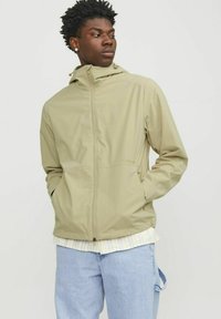 Lys beige vandtæt jakke med lynlås, hætte og to frontlommer. Har en glat tekstur og et minimalistisk design.