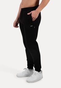 Sorte sweatpants lavet af blødt stof med elastisk talje, sidelommer og tætsiddende design, parret med hvide sneakers.
