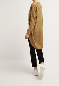 Cardigan long et ample en tricot doré métallique, avec un dos drapé. Associé à un pantalon noir et des baskets blanches.