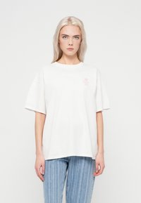Hvid bomulds t-shirt med korte ærmer, rund hals og et lille pink blomstermotiv på venstre bryst. Båret med lyseblå stribede jeans.