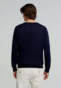 Pulover tricotat de culoare albastru închis, cu guler rotund, mâneci lungi și manșete cu nervuri, purtat cu pantaloni de culoare deschisă, evidențiind o textură fină.