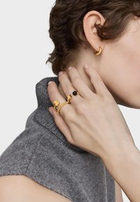 Pendientes de oro y varios anillos de oro con una piedra negra. La mano reposa sobre un suéter gris texturizado.