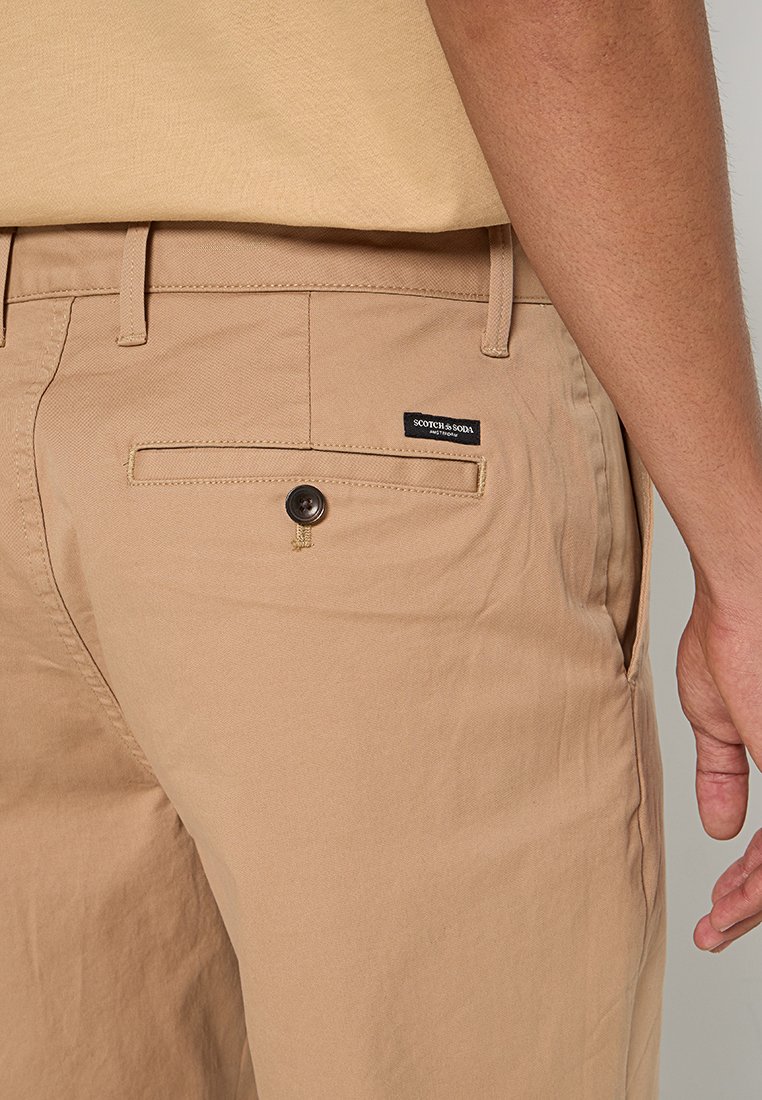 Beige Baumwollhose mit einer Gesäßtasche, die mit einem Knopfverschluss ausgestattet ist, sowie einem kleinen Markenlabel. Der Stoff hat eine glatte Textur.
