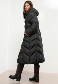 Cappotto lungo nero di piumino con cappuccio, motivo a zig-zag, tasche laterali e dettaglio in vita arricciato, abbinato a stivali neri al ginocchio.
