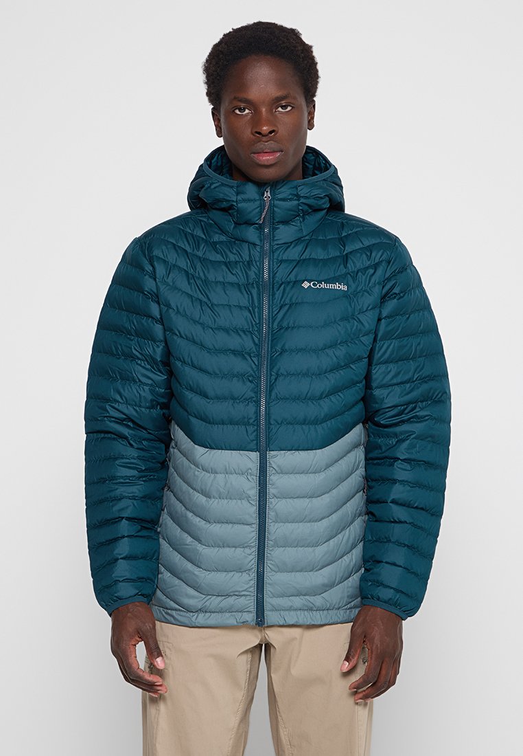 Hombre con chaqueta acolchada con capucha de Columbia en tonos verde azulado y azul claro, cerrada, de pie frente a un fondo gris liso.