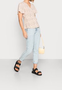 Blusa de color rosa claro con escote en V y mangas cortas con volantes, con un patrón oscuro disperso, combinada con jeans azul claro enrollados y sandalias negras.