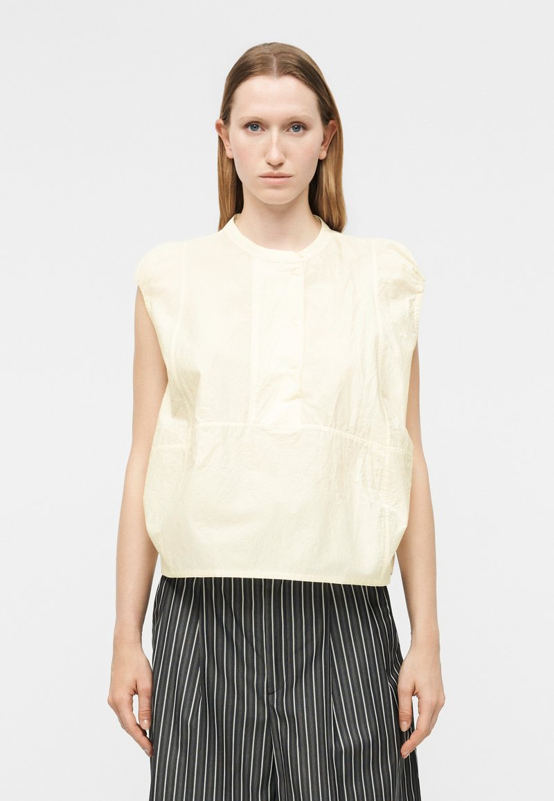 Γυναίκα με ίσια ανοιχτόκαστα μαλλιά φοράει αμάνικο κρεμ τοπ και μαύρο παντελόνι με λευκές ρίγες, στέκεται μπροστά σε απλό φόντο.