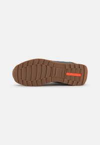 Sole di sneaker nera con un battistrada in gomma marrone testurizzata, dotata di una placca logo arancione rettangolare. Progettata per la trazione e la durata.