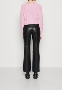 Suéter cortado de punto trenzado en color rosa, combinado con pantalones de cuero sintético negros de tiro alto y piernas anchas, y botas gruesas negras, visto desde atrás.