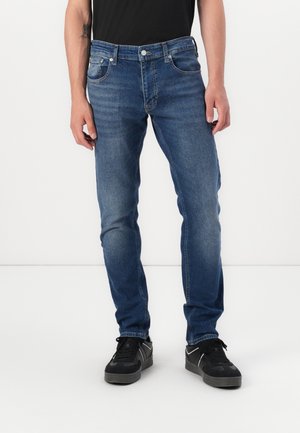 Homme portant un jean bleu slim avec des détails délavés et des baskets noires, debout devant un fond blanc uni.