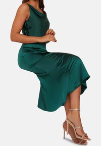 Smaragdově zelené saténové šaty s kulatým výstřihem a rozšířeným lemem, doplněné o nahé sandály na vysokém podpatku se zdobením z rhinestones.