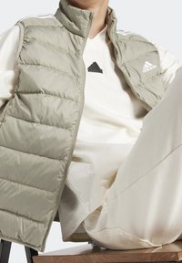 Světle zelená péřová vesta s vysokým límcem, zipovým zapínáním a horizontálním prošíváním, nošená přes krémovou košili s logem.