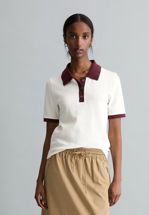 Młoda kobieta stojąca, ubrana w białą koszulkę polo z krótkim rękawem, z ciemnoczerwonym kołnierzem i mankietami oraz beżową spódnicę z troczkami i kieszeniami na zamek.