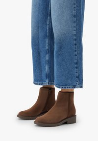 Una persona vistiendo jeans azules recortados y botas de ante marrón hasta el tobillo con cremalleras laterales y tacones bajos, de pie sobre una superficie blanca.