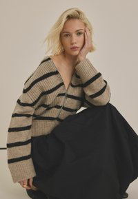 Beige och svart randig stickad tröja med dragkedja framtill, ribbad textur och vida ärmar, ihop med en svart kjol.