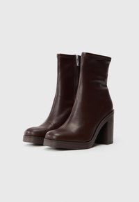Botine din piele maro, cu toc bloc înalt, textură netedă, vârf rotund și detaliu cu fermoar pe lateral. Design simplu și modern.