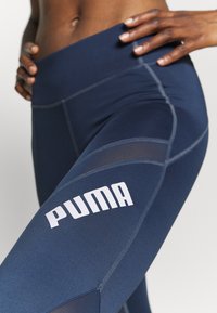 Marinblå sportleggings med hög midja, prydda med vit "PUMA"-logotyp och kontrasterande strukturerade paneler för flexibilitet och stil.