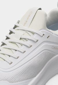 Άσπρο αθλητικό παπούτσι Calvin Klein με πλέγμα στα πάνελ, παχιά σόλα και άσπρα κορδόνια, που δείχνει κοντινή εικόνα της πλαϊνής πλευράς και της ετικέτας στη γλώσσα.