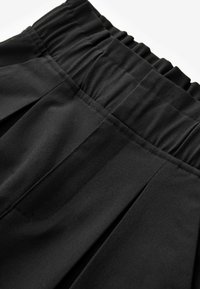 Pantalon noir avec taille élastique et coutures verticales visibles, mettant en valeur la texture du tissu et les détails de la couture.