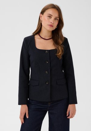 Jonge vrouw met lang bruin haar die een donker krijtstreepblazer met grote knopen en een donkere spijkerbroek draagt tegen een effen achtergrond.