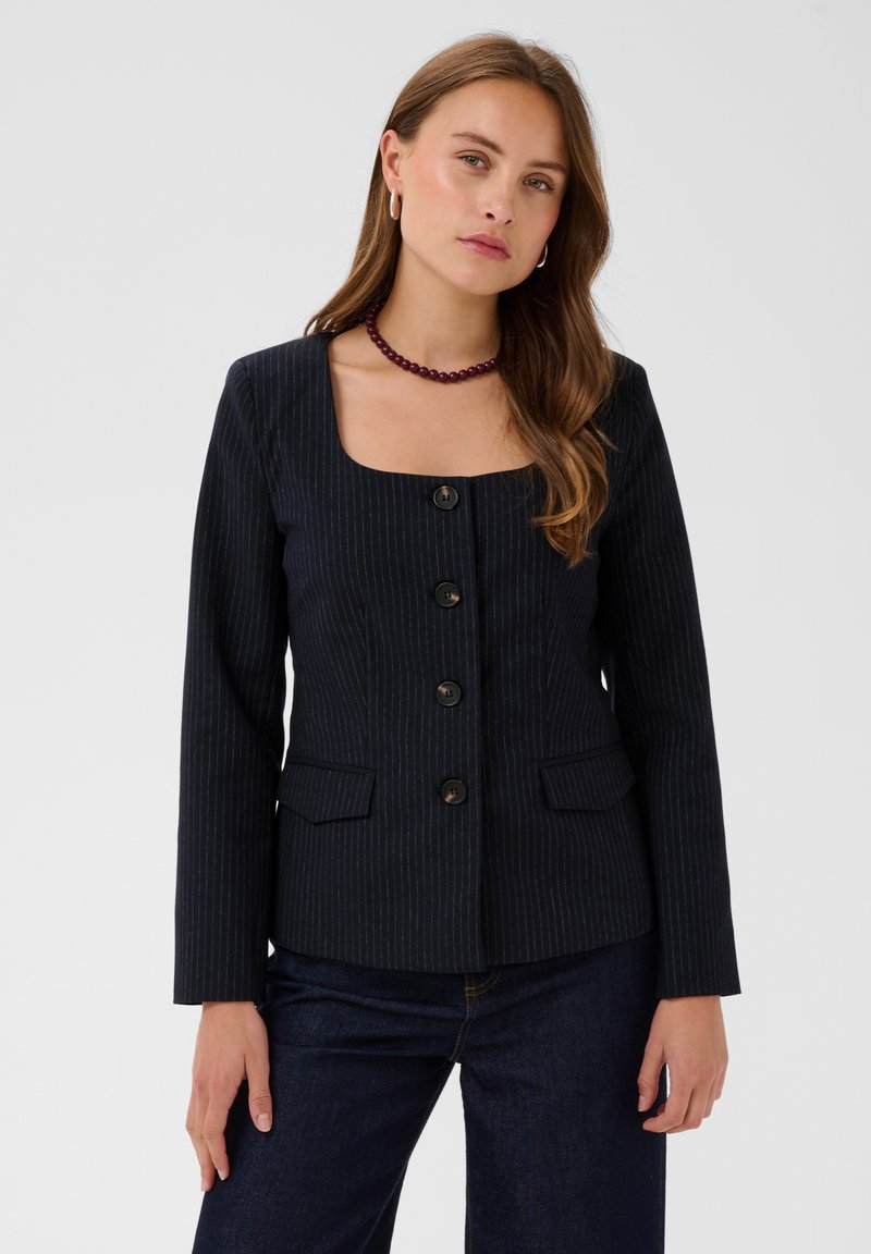 Jeune femme aux longs cheveux bruns portant un blazer sombre à fines rayures avec de gros boutons et un jean foncé sur un fond uni.