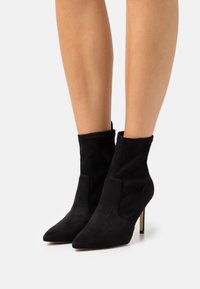 Pair of black suede pointed-toe ankle boots with stiletto heels worn on bare legs against a plain background.