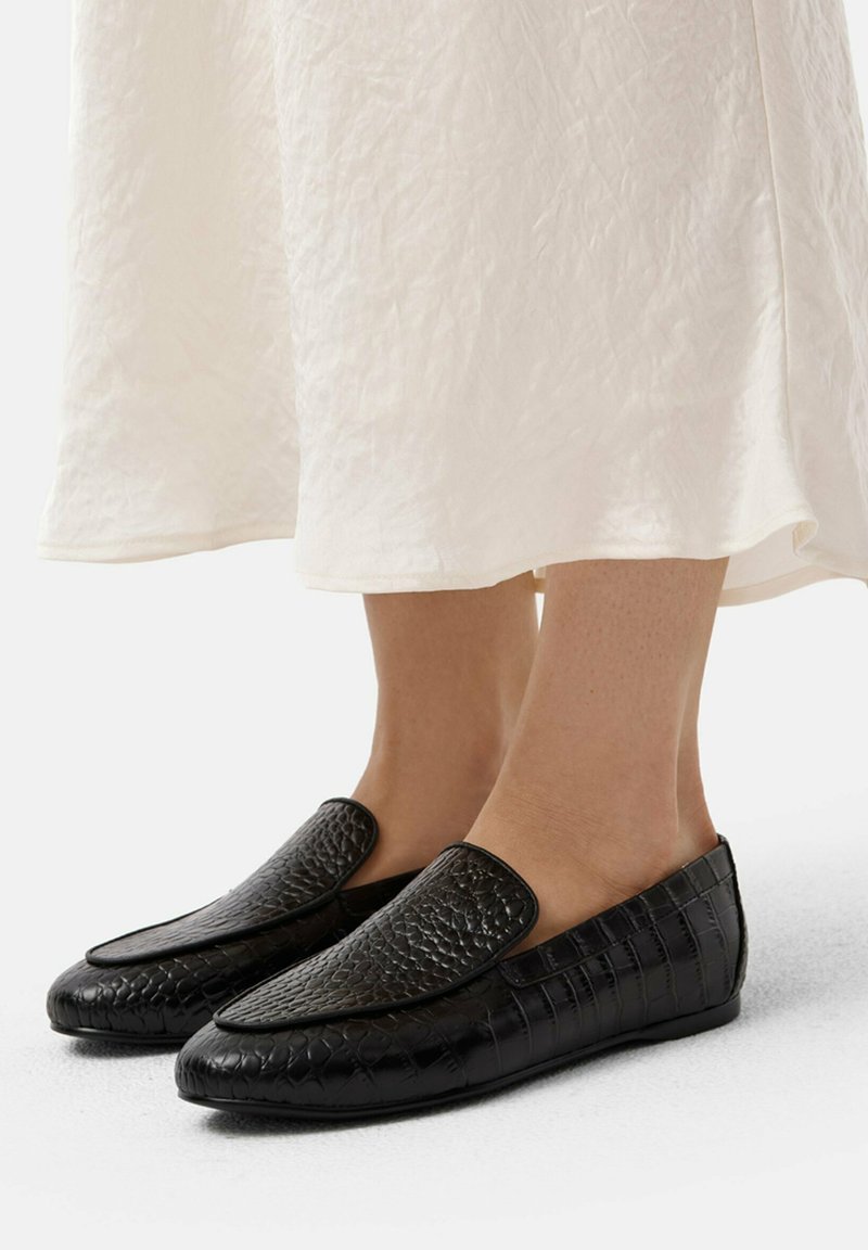 Zwarte instapschoenen hebben een gestructureerd, reptielachtig patroon aan de voorkant met een soepele, platte zool. Draagt met een lichtgekleurde jurk.