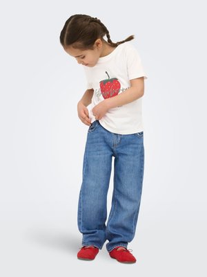 Dijete s pletenicama koje nosi bijelu majicu s printom jagoda, široke plave traperice i crvene cipele, gleda dolje dok prilagođava džep na trapericama.