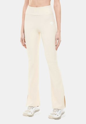 Donna che indossa pantaloni beige a zampa con spacchi laterali e sneakers bianche firmate EA7, in piedi davanti a uno sfondo semplice.