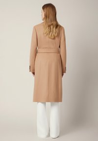 Karmelowy długi płaszcz o strukturalnym designie, z paskiem w talii i zapięciem na guziki przy mankietach. Materiał wydaje się teksturowany i odpowiedni na chłodniejsze dni.