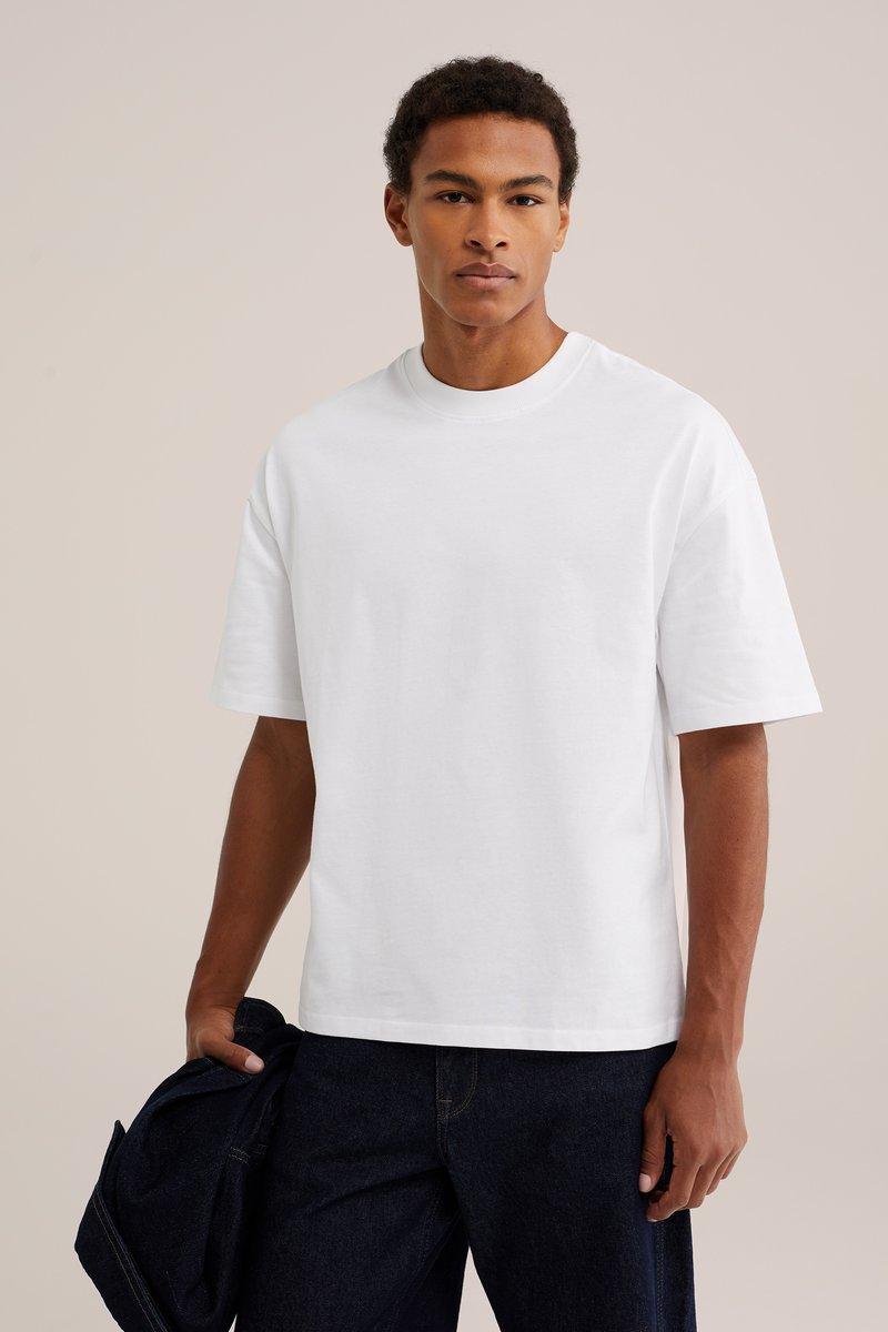 Jeune homme portant un t-shirt blanc uni à manches courtes et un pantalon sombre, tenant une veste sombre dans sa main gauche devant un fond uni.